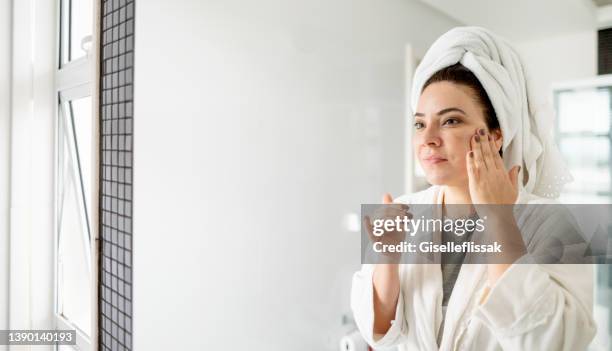 woman wearing a towel and bathrobe applying moisturizer to her face - cream for face stock pictures, royalty-free photos & images