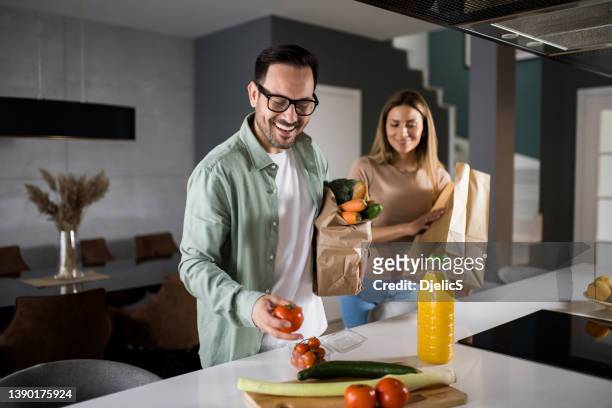 happy couple returning home from shopping trip and unpacking grocery bags - young man groceries kitchen stock pictures, royalty-free photos & images