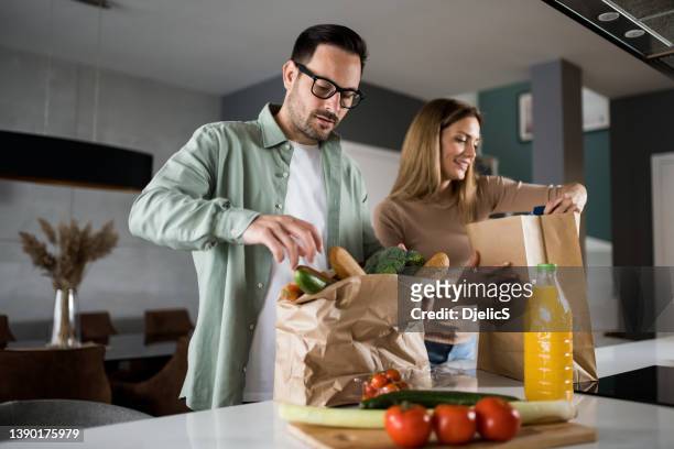 couple returning home from shopping trip and unpacking grocery bags - young man groceries kitchen stock pictures, royalty-free photos & images