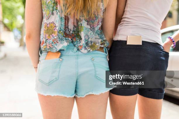 teenage girls standing with smart phones in pockets on sunny day - hot pants stock pictures, royalty-free photos & images