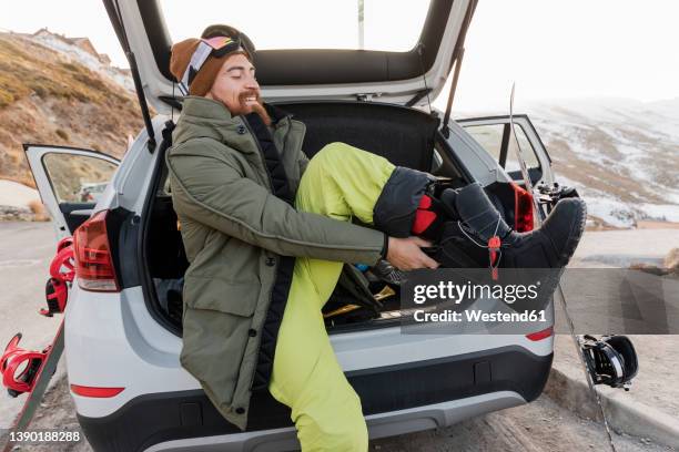 smiling young man in car trunk putting on ski boots - skischoen stockfoto's en -beelden