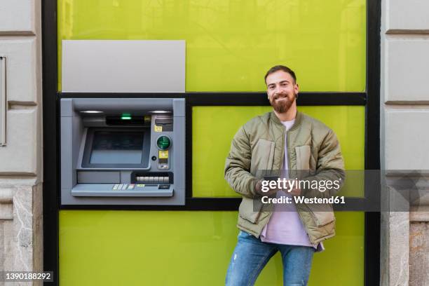 smiling young man with smart phone standing by atm - man atm smile stock pictures, royalty-free photos & images