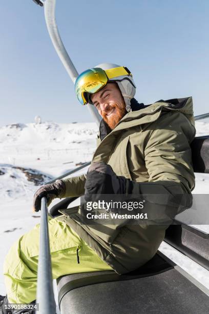 happy young man gesturing with closed fist sitting on ski lift - ski jacket stock pictures, royalty-free photos & images