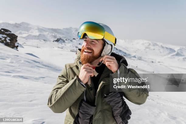 smiling young man with beard fastening skiing helmet - skikleidung stock-fotos und bilder