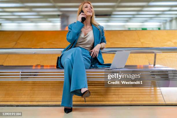 smiling blond businesswoman talking on mobile phone sitting with laptop on bench at subway station - mujeres de mediana edad fotografías e imágenes de stock