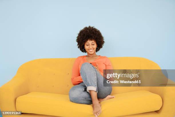 happy young woman surfing net through smart phone sitting on yellow sofa against blue background - sitting and using smartphone studio stock pictures, royalty-free photos & images