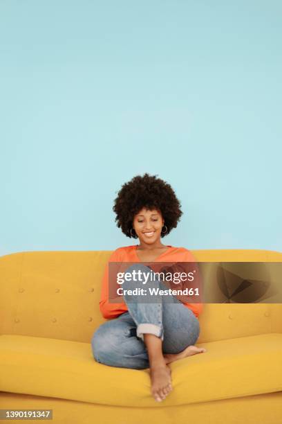 happy woman using mobile phone sitting on yellow sofa against blue background - woman on phone isolated stock pictures, royalty-free photos & images
