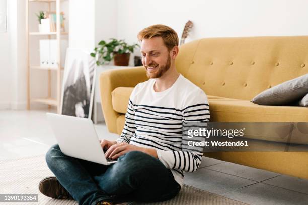 smiling young man using laptop sitting cross-legged in front of sofa at home - person in front of computer stock pictures, royalty-free photos & images
