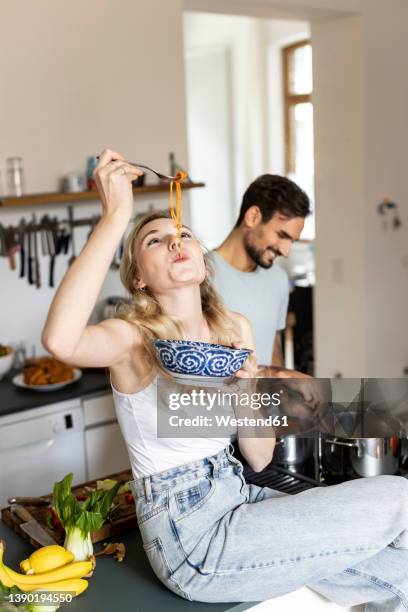 woman slurping noodles with boyfriend preparing food in kitchen at home - couple in kitchen foto e immagini stock