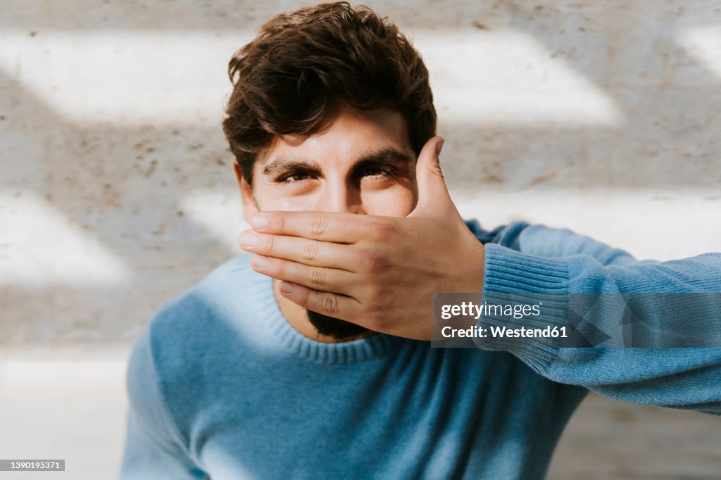 Young man covering mouth with hand standing in front of wall