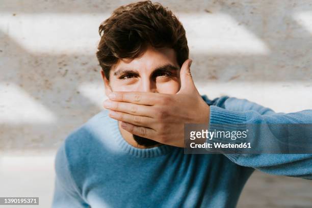 young man covering mouth with hand standing in front of wall - hands covering mouth stock pictures, royalty-free photos & images