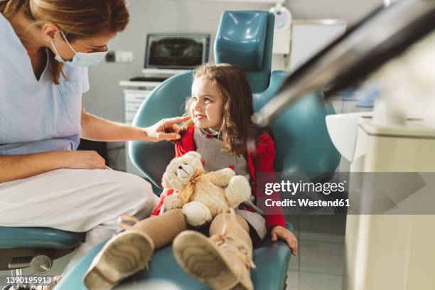 dentist in protective face mask looking at girl sitting with stuffed toy in dental clinic - 小児歯科 ストックフォトと画像