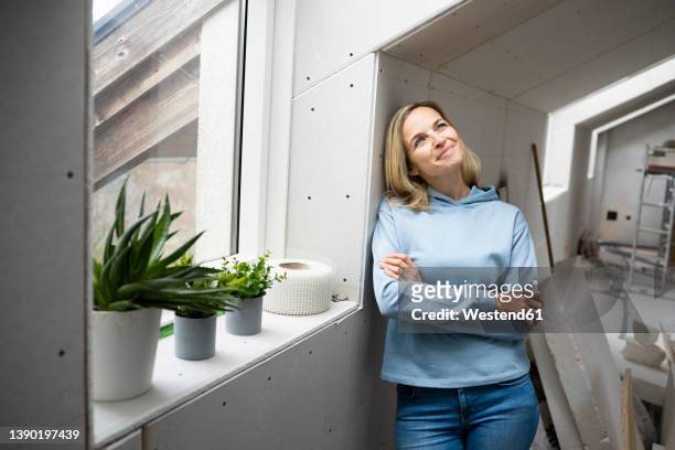 thoughtful woman with arms crossed standing by window in attic - combles photos et images de collection