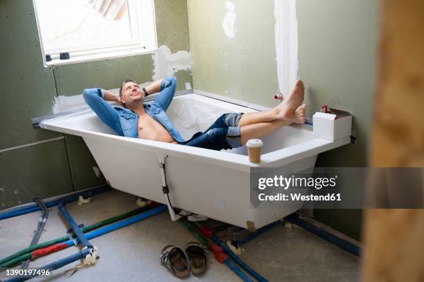 smiling man with hands behind head lying in bathtub in attic - unvollständig stock-fotos und bilder
