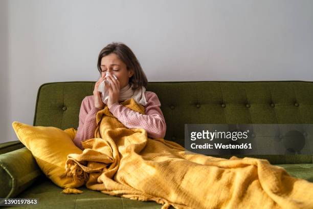 sick woman blowing nose in facial tissue at home - flu stockfoto's en -beelden