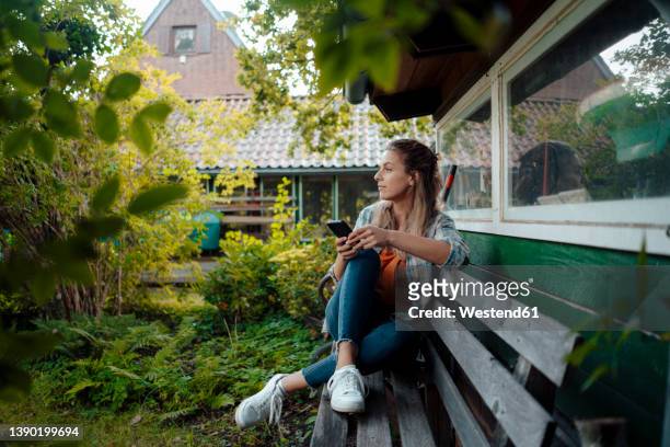 woman with smart phone sitting on bench at backyard - garden bench stock pictures, royalty-free photos & images