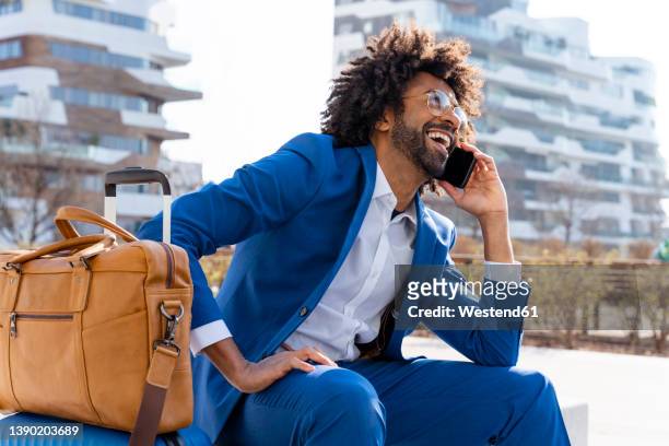 happy businessman with suitcase and shoulder bag talking on smart phone sitting on concrete block - viagem de negócios imagens e fotografias de stock