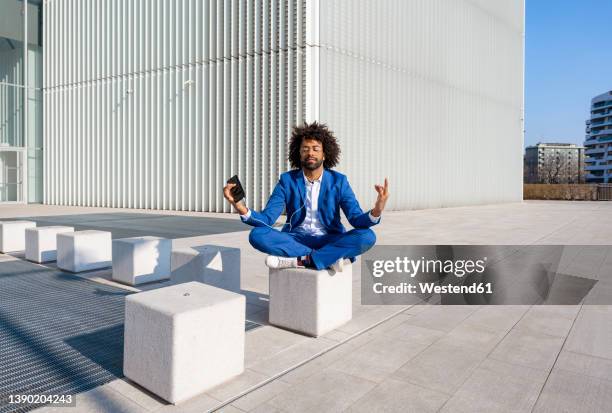 businessman with mobile phone listening music and meditating on concrete block - businessman meditating stock pictures, royalty-free photos & images