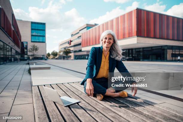 happy businesswoman sitting with cross-legged by laptop at office park - tailleur photos et images de collection