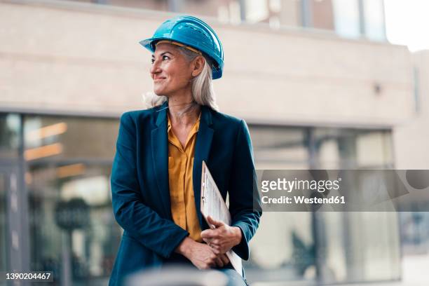 smiling engineer wearing hardhat holding clipboard at office park - ingenieur stock-fotos und bilder