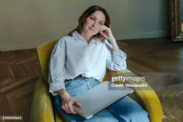 thoughtful woman with laptop sitting on yellow armchair - donna poltrona foto e immagini stock