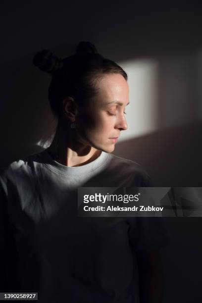 a lonely young sad calm girl or woman stands against a white wall, in a psychiatric clinic or at home, in the rays of sunlight, with her eyes closed. the concept of depression from illness and quarantine, fatigue and impotence. a copy of the space. - woman prison stock-fotos und bilder