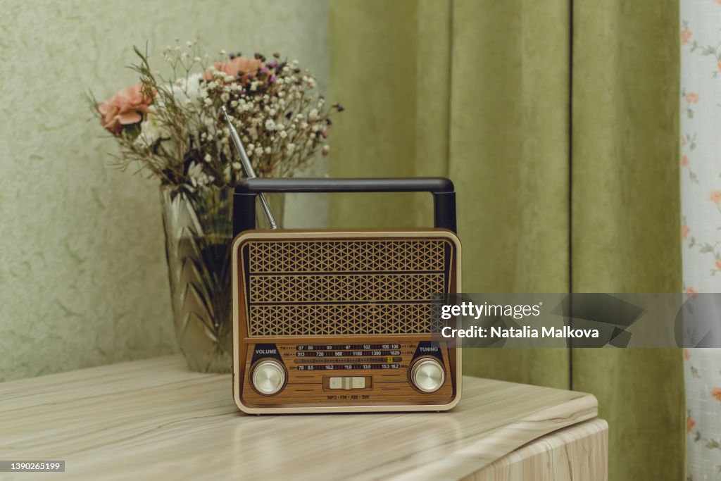 On a light wooden chest of drawers near the window with light green curtains, there is a radio receiver in an old retro style, next to it is a glass vase with various flowers, copy space.