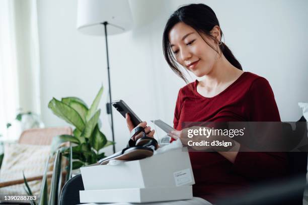 smiling young asian woman with smartphone, receiving packages that she have purchased from shopping online and unboxing to check the products at home. convenience and easy online shopping experience. contactless delivery service makes life so much easier - home delivery stock pictures, royalty-free photos & images