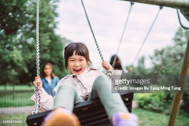 lovely little girl smiling at the camera while playing on a swing set in playground joyfully - playing imagens e fotografias de stock