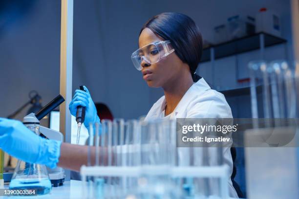 una joven, hermosa y enfocada trabajadora de laboratorio con gafas protectoras en su escritorio con matraces de laboratorio y tubos de ensayo de vidrio en un estante de tubos de ensayo - secuenciación de polímeros fotografías e imágenes de stock