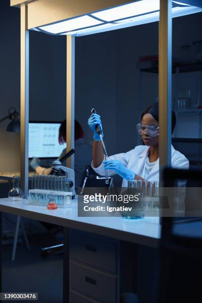 schöne afroamerikanische laborärztin bei der arbeit, sitzt an ihrem schreibtisch in einem modernen labor und mischt flüssigkeiten in einem reagenzglas - genome sequencing stock-fotos und bilder