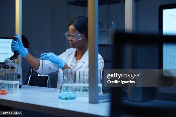 eine afroamerikanische wissenschaftlerin bei der arbeit, die an ihrem schreibtisch in einem modernen labor sitzt und chemische reaktionen in einem reagenzglas durchführt - biotechnology lab stock-fotos und bilder