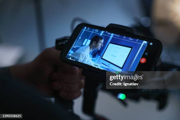 a backstage from the set and a smartphone in a director's hand with the image of a scientist working in front of a computer monitor - film set stockfoto's en -beelden