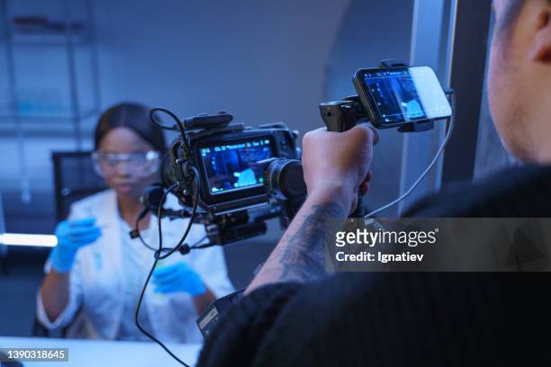 eine professionelle kamera, die eine afroamerikanische schauspielerin in der rolle eines wissenschaftlers in einem labor fotografiert. backstage vom filmen eines fotosets in einer labordekoration und einer bedienerschulter im vordergrund - filmregisseur stock-fotos und bilder
