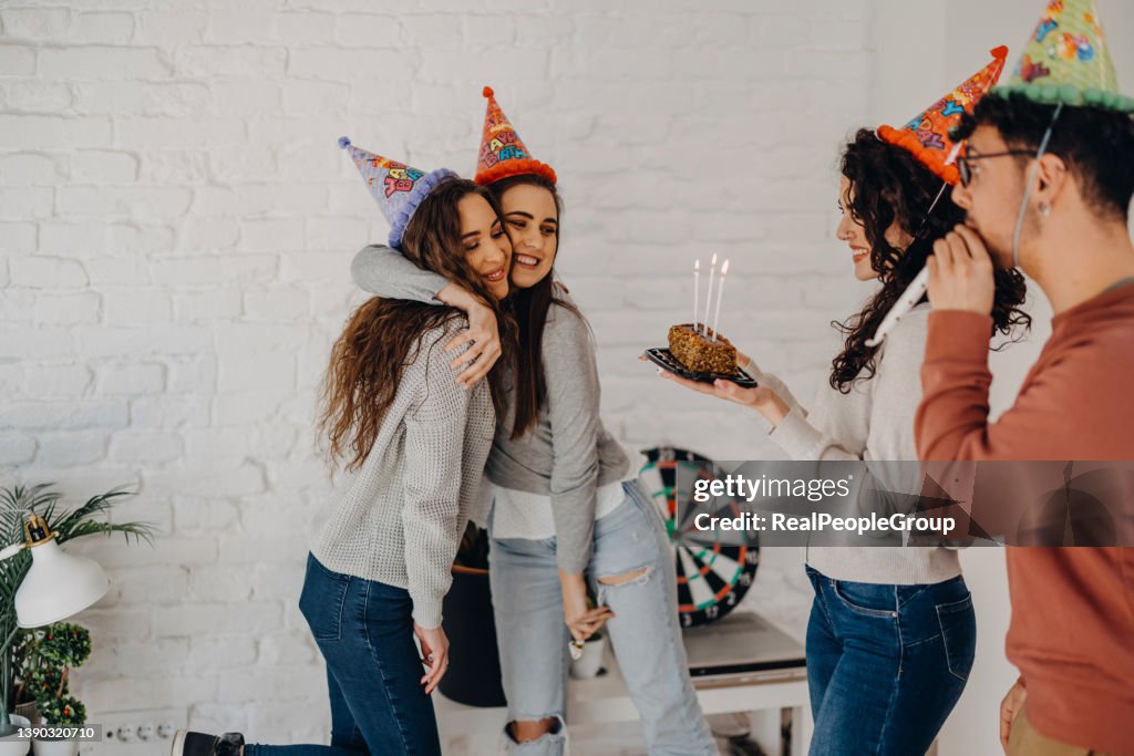 Young Friends making surprise for birthday woman, holding cake with lit candles, wearing party hats and smiling