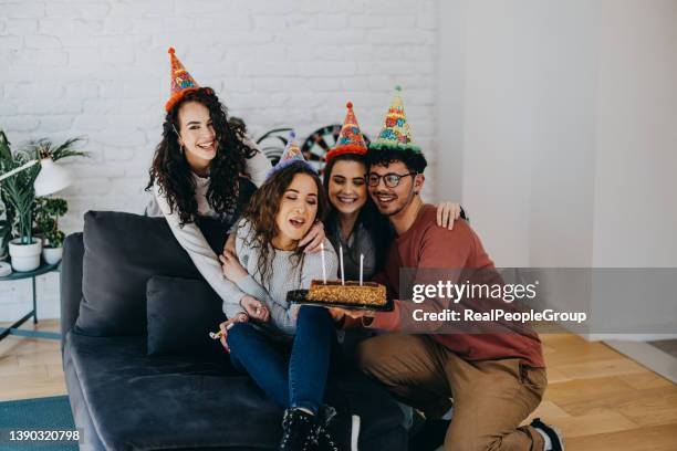 young friends making surprise for birthday woman, holding cake with lit candles, wearing party hats and smiling - college dorm party stock pictures, royalty-free photos & images