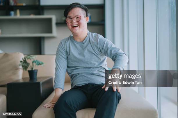 porträt asiatisches chinesisches down-syndrom junger mann schaut lächelnd in die kamera im wohnzimmer - downsyndrom stock-fotos und bilder