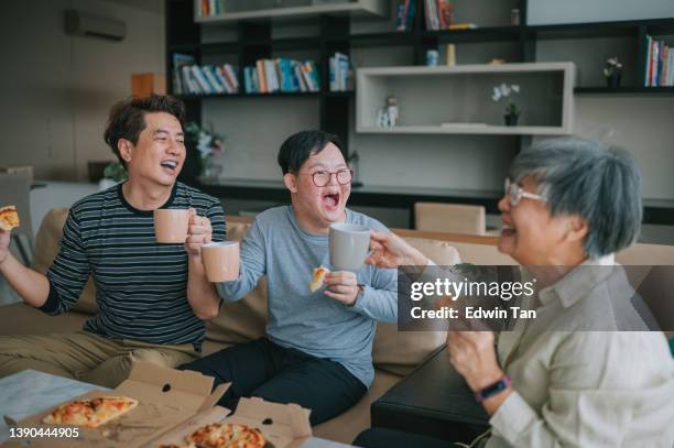 asiatische chinesische mehrgenerationenfamilie mit down-syndrom-enkel, der pizza fast food im wohnzimmer genießt und am wochenende auf dem sofa sitzt, mit feiertoast - family on couch with mugs stock-fotos und bilder