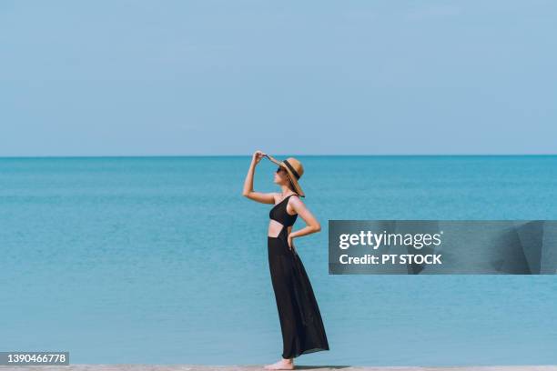 a woman in a black bikini, wearing a long black skirt and wearing a hat, stands by the sea and the sky is blue. - luxury hotel island stockfoto's en -beelden