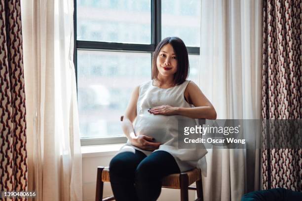 asian pregnant woman rubbing her belly while sitting by the window enjoying warm sunlight. - birthing chair stock pictures, royalty-free photos & images