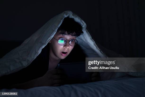 cute girl watching video on phone with shocked facial expression in bed at night - horror room stock pictures, royalty-free photos & images