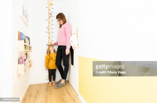 front view of little girl looking up at her mother with a big smile as her mother measures her, together in a small children's corner. - measure length stock pictures, royalty-free photos & images