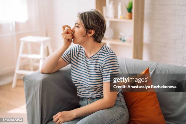 woman sits on couch and inhales medicine from an inhalation can - asmático fotografías e imágenes de stock