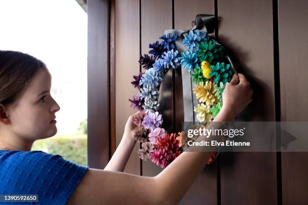 teenage girl positioning a multi-colored homemade decorative wreath made from painted pine cones, onto a front door at easter time. - wreath making stock pictures, royalty-free photos & images