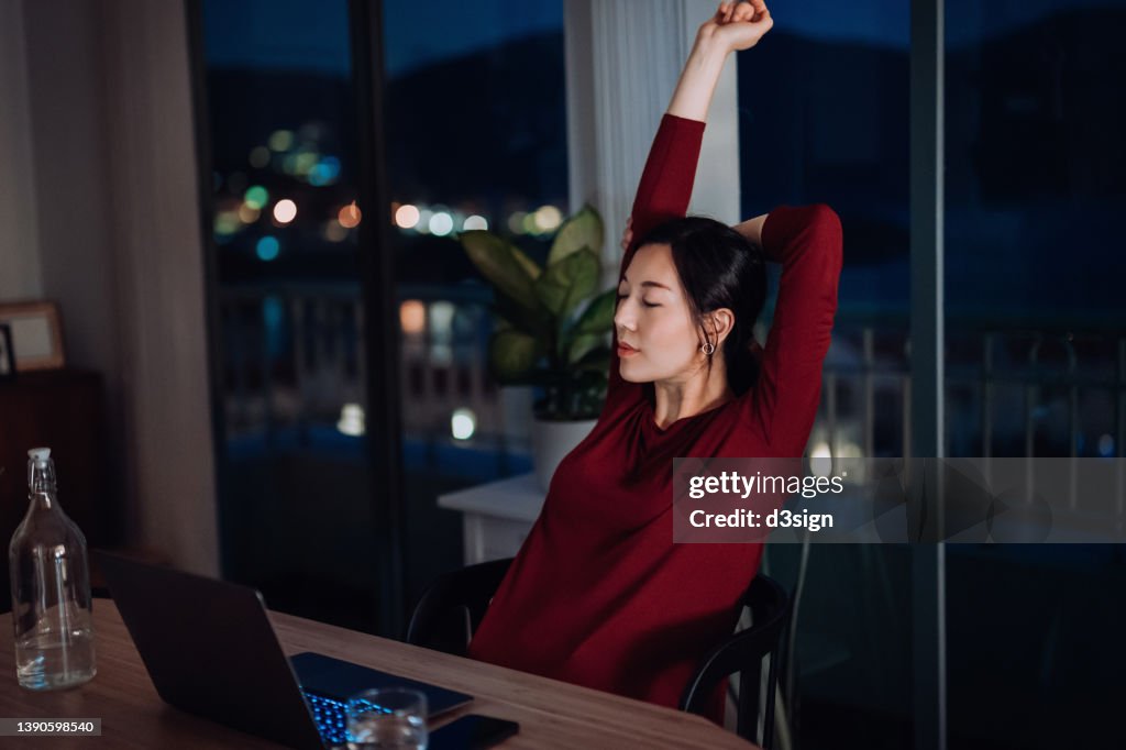 Exhausted young Asian businesswoman stretching with arms raised while taking a break at her desk working late in home office at night
