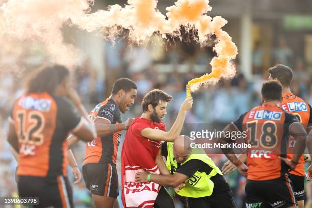 Joe Ofahengaue of the Tigers holds a pitch invader carrying a flare as he is tackled by security during the round five NRL match between the Cronulla...