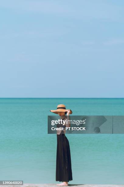 a woman in a black bikini, wearing a long black skirt and wearing a hat, stands by the sea and the sky is blue. - long skinny legs foto e immagini stock