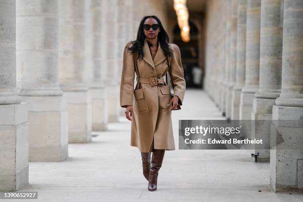 Emilie Joseph @in_fashionwetrust wears black sunglasses, rhinestones earrings, a black shirt, a beige long belted trench coat, a burgundy shiny...