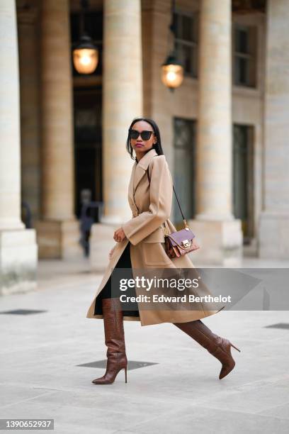 Emilie Joseph @in_fashionwetrust wears black sunglasses, rhinestones earrings, a black shirt, a beige long belted trench coat, a burgundy shiny...