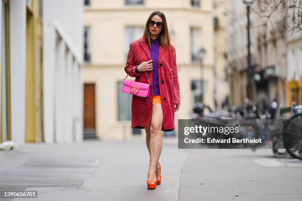 Maria Rosaria Rizzo @lacoquetteitalienne wears white pearl pendant earrings, a purple shirt, a neon orange short skirt, a red velvet long coat, a...